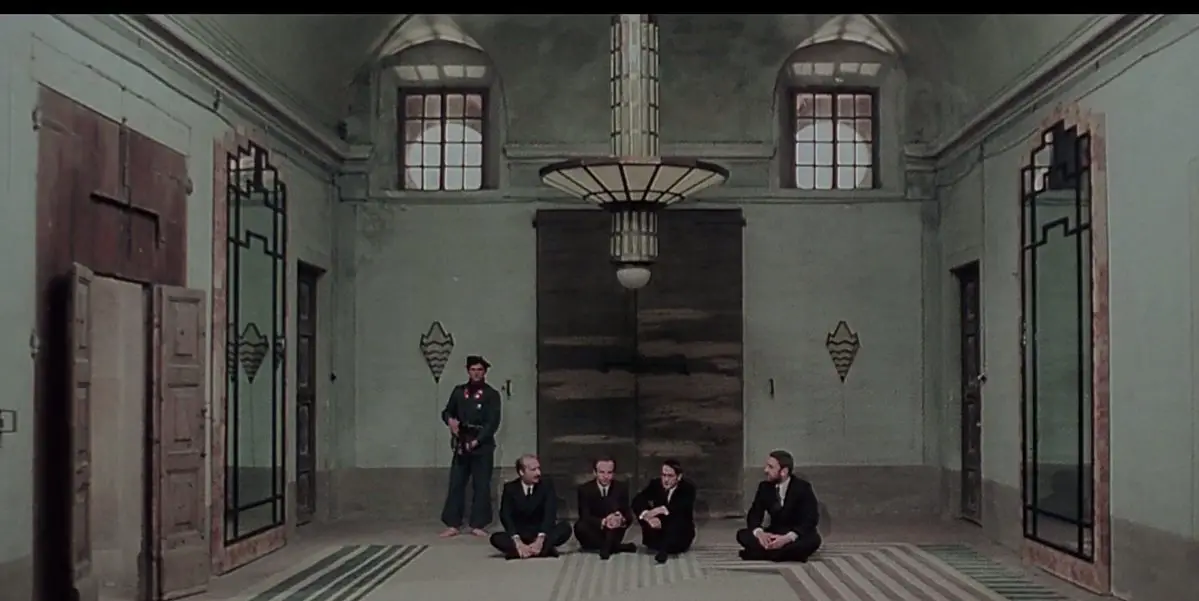 Four young boys sit and wait on a tile floor in a large room