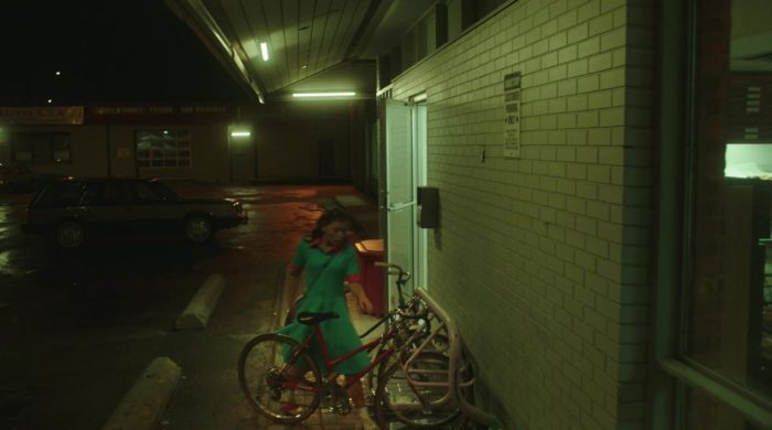 A young woman is about to get on her bike in a dark, deserted parking lot at night.
