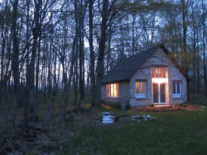 A cabin sits in the middle of the woods lit during dusk.