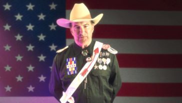Joe Bob Briggs, standing in front of an American flag