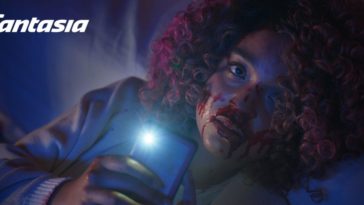 Cecilia sits, her face covered in blood, aiming her phone's flashlight toward the camera in Sissy