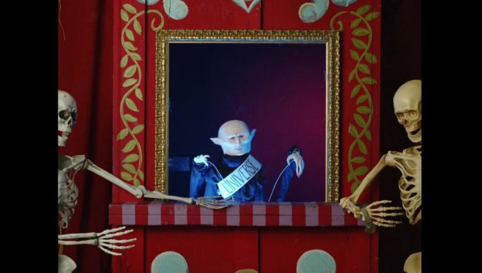a vampire puppet wearing a Mr. Universe sash appears between two skeletons