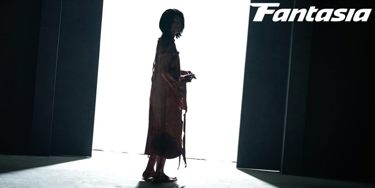 Girl stands wearing a bloody medical gown in a large doorway letting light into a dark room in The Witch Part 2: The Other One