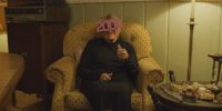 An old woman sits in a chair wearing party glasses bearing the year 2000 on them while giving a thumbs up in V/H/S/99