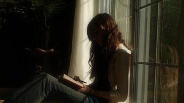 A person with long hair reads a book while sitting in shadows illuminated by a sunny window.