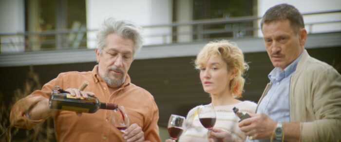 Jeanne and Gérard hold champagne glasses to the right of Alain who is pouring another glass in Incredible but True