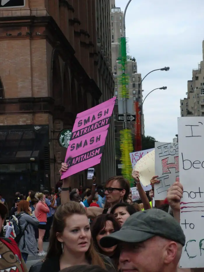 A crowd in a protest, one person holding a sign saying 'SMASH PATRIARCHY SMASH CAPITALISM'