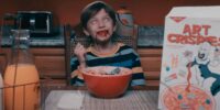 A possessed-looking smiling child bleeding from the mouth holding a spoon over a bowl of cereal with something undeterminable in the bowl next to a cereal box for Art Crispies in the Terrifier 2 trailer