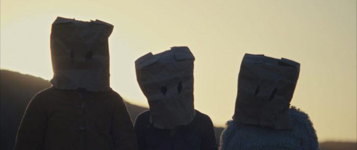 Three people stand side by side wearing paper bags over their heads with the eyeholes cut out