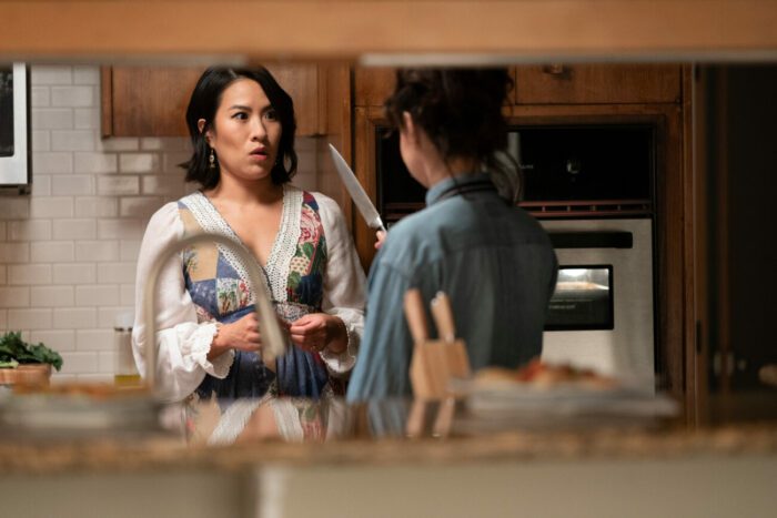 A woman holds a knife up to Margo in the kitchen 