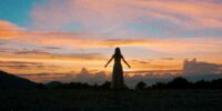A woman stands, arms outstretched, looking out at a sunset