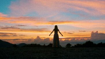 A woman stands, arms outstretched, looking out at a sunset