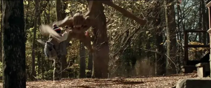 A young child standing in the woods is about to be attacked by a large monster. The monster is brown with long arms and lungs and a sharp ending at the end of each arm. It is jumping through the trees towards the child.
