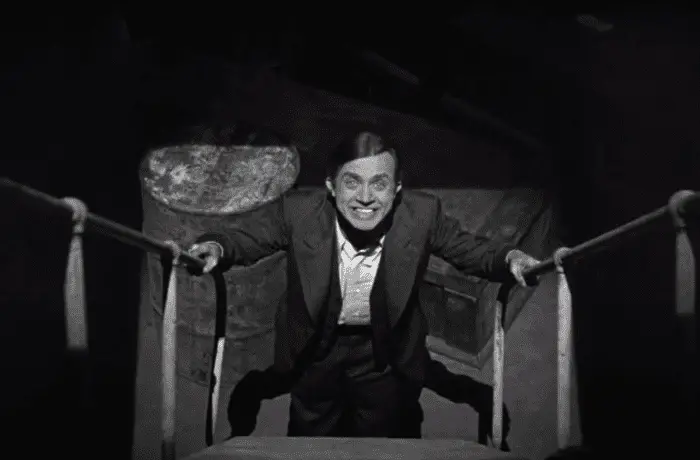 Renfield stands at the bottom of the stairs, completely insane, grinning wildly up at the camera in Bela Lugosi's Dracula