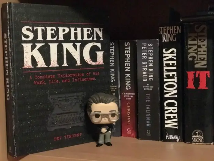 A Stephen King doll stands on a bookshelf next to Stephen King books and The Stephen King Ultimate Companion by Bev Vincent