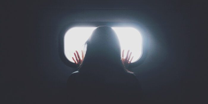 In darkness a long haired person looks out of a rectangular window with their hands pressed against the glass. Beyond, a white light bursts through, illuminating their hands.