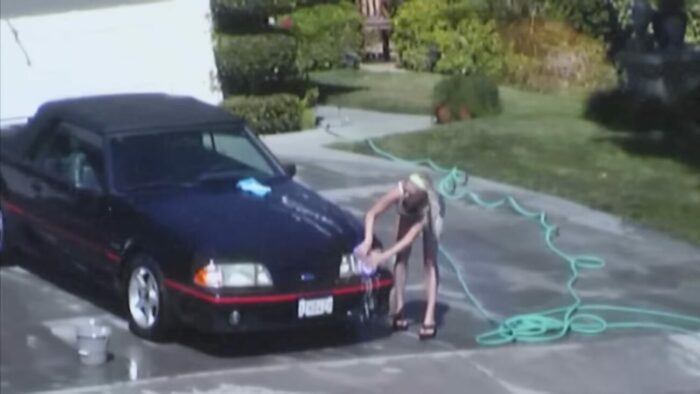 A woman is washing her car as she is filmed by the kids across the street