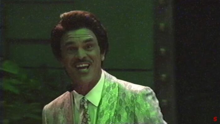 The sleazy and creepy gameshow host dons a toothy grin while bathed in green light