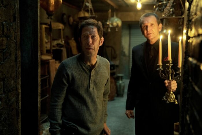 Two men go deeper in a packed storage unit, one holding a candelabra 