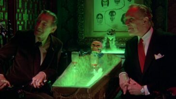 David Carradine and Vincent Price muse about something happening off screen with a glass-covered, neon green lit coffin acts as a table between them in The Monster Club
