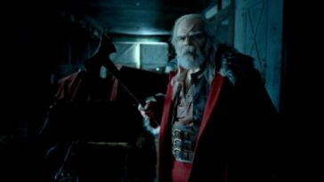 Santa is standing in a stable holding an ax in front of his sleigh with blood streaking down his forehead and nose in A Christmas Horror Story