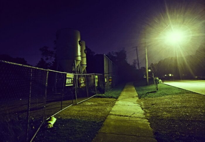 An empty suburban street at night lit by a gleaming streetlamp shining on a dark, vacant construction lot