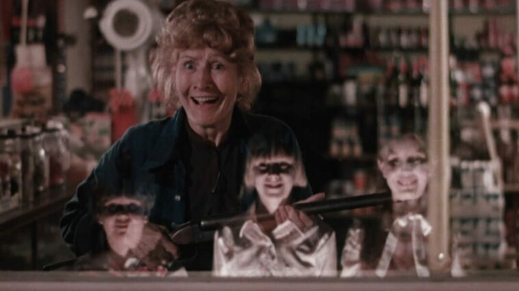three devilish looking children are reflected in a window where, on the other side, a woman holds a shotgun.