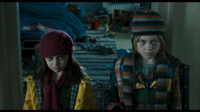 Two children shand in front of a messy play room in winter hats, scarves, and jackets