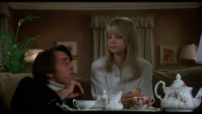 A young girl and looks down at an older gentleman sitting below her as they share afternoon tea.