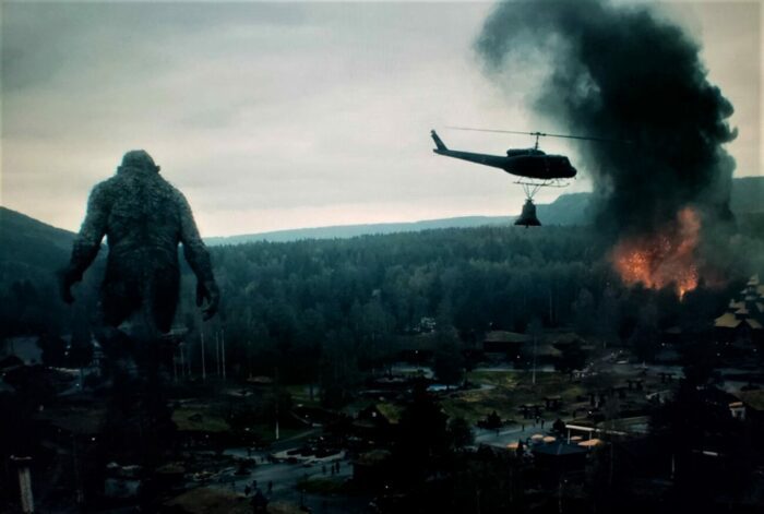 Helicopter carrying a bell circles a colossal troll as a city below burns.