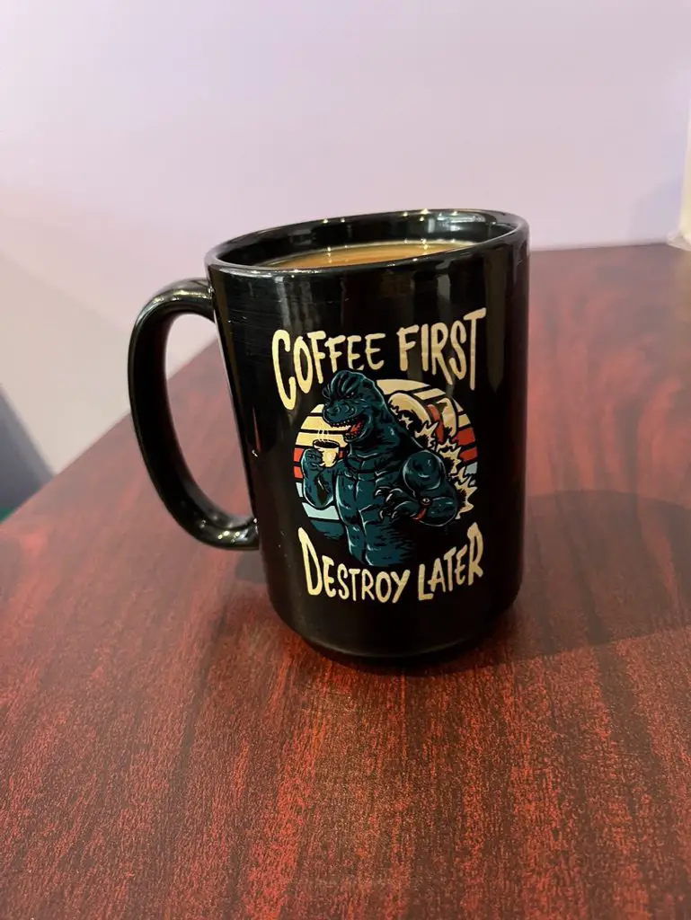 A black mug full of coffee with a cartoon of a green Godzilla with the words "Coffee First Destroy Later," at the coffee shop, "The Brewed."
