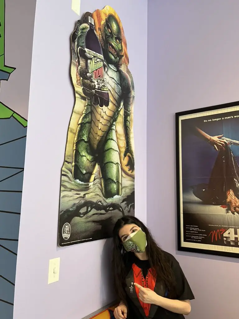 Jamie Lee, in a Svengoolie costume T-shirt, sits in front of and points to a cardboard standee on the wall of the Creature from the Black Lagoon holding a bottle of Mountain Dew Pitch Black at the coffee shop, "The Brewed."
