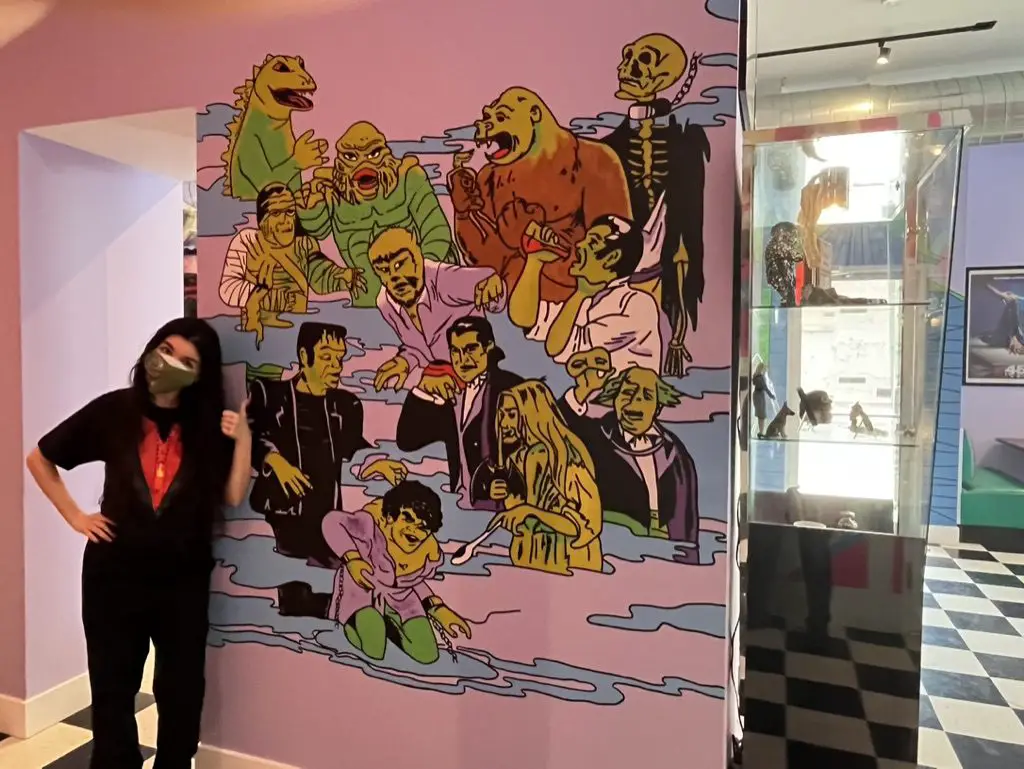 Jamie Lee, in a Svengoolie costume T-shirt, stands in front of and points to a mural of various Universal Monsters and other movie monsters, painted by Jen Lemasters, at the coffee shop, "The Brewed."