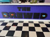 The logo for coffee shop "The Brewed" against purple bricks on the front of the front counter.
