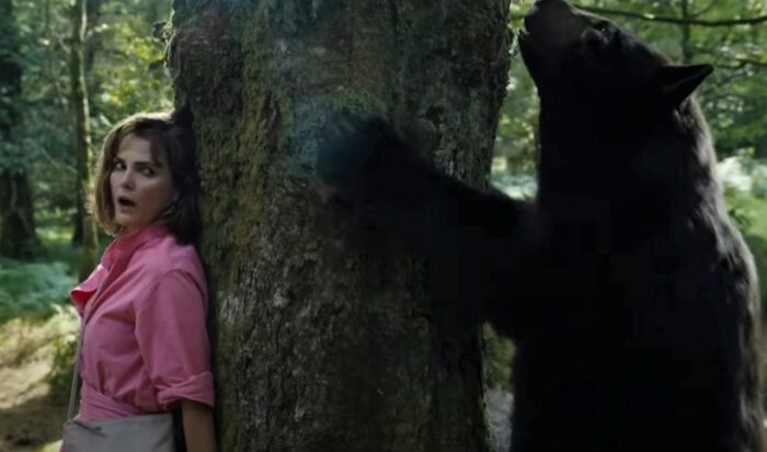 Keri Russell hides behind a tree as the titular Cocaine Bear claws at the trunk on the opposite side.