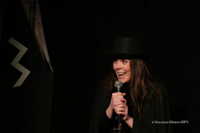 Maria stands center stage, wearing a top hat, plastic fangs, and a black cape