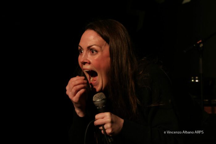 Maria stands center stage, yelling towards the audience with a microphone in her left hand