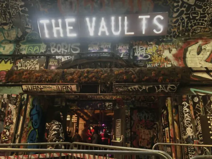 Front entrance to The Vaults, inset in the tunnel, covered in graffiti, with bright white lights spelling The Vaults.