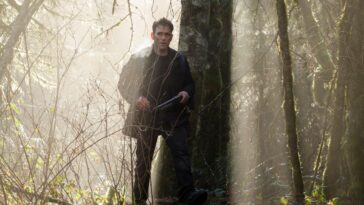 Ethan Burke stands in the forest next to a large tree with a weapon in his hand. Rays of sunshine pour down around him