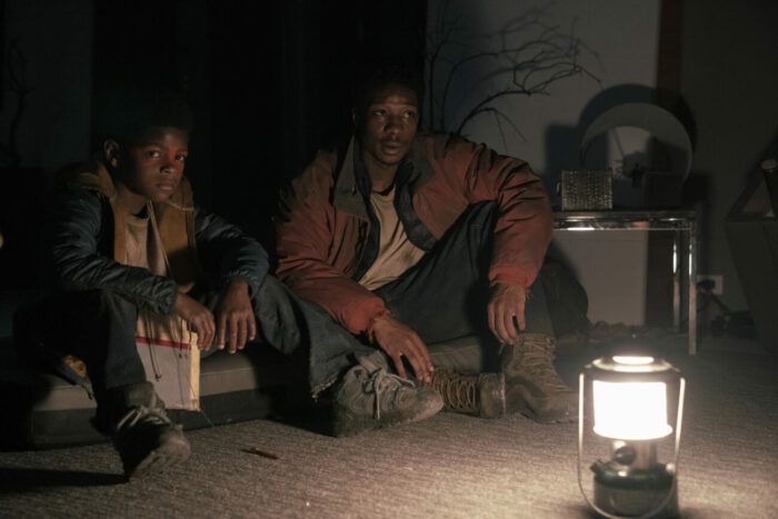 Sam and Henry sit together in the glow of lantern light in The Last of Us S1E5 "Endure and Survive"