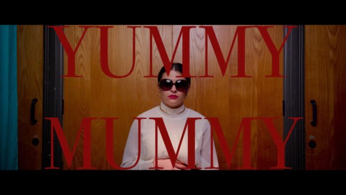 Title card for Yummy Mummy, red text spells "Yummy Mummy" overlayed on a still of Lilith at the doctor's office.