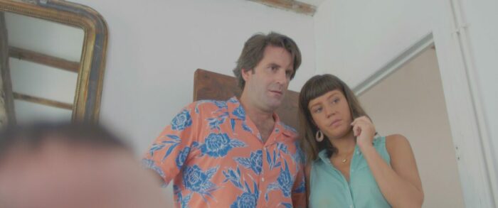 A man in a bright floral shirt speaks to a woman in a sleeveless bright teal blouse standing beside him.