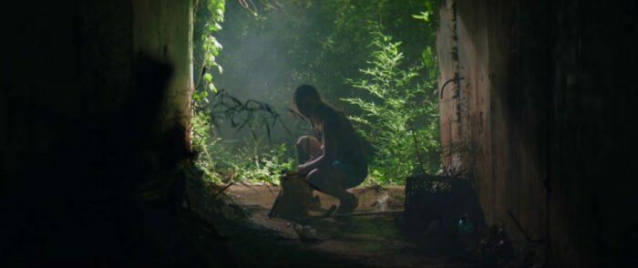August kneels down in a cavern leading out into some brush