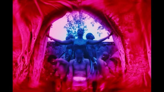 From Ni'jah's music video Festival, three people are on their knees in front of Ni'jah, with two people behind her. Her arms are raised to her shoulders. The colors are cosmically red and purple