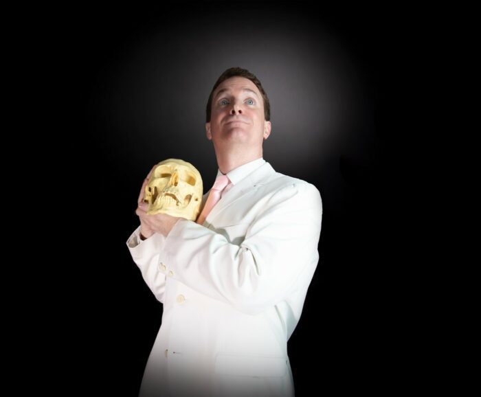 Grady Hendrix stands in a white suite and pink tie holding a skull. Hendriv will attend CFF23.