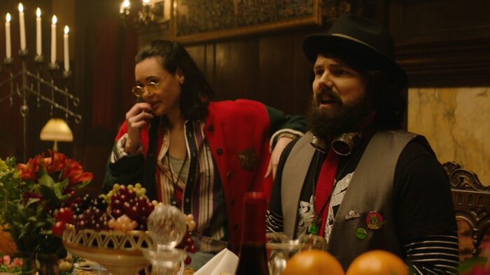 A woman in a red jacket and glasses presses her fingers to her lips next to a bearded man in a black top hat and grey vest, both sit in front table covered in food.