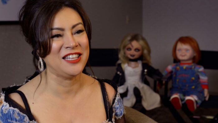 Jennifer Tilly smiles while dolls Tiffany and Chucky wach her in the background in Living with Chucky