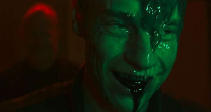 A smiling green-hued man with blood dripping from his mouth is covered in slime 