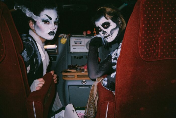 two people in the front seat of a car turned to face the camera in the backseat. They are wearing face paint and resemble The Bride of Frankenstein and a skeleton