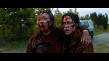 Two bloodied and exhausted people embrace while staring off into the distance.